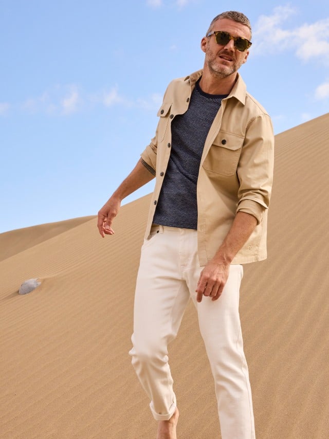 Ein Mann steht in den Dünen und trägt eine helle Hemdjacke, darunter ein Shirt und dazu eine weiße Hose. Ein Mann steht in den Dünen und trägt eine helle Hemdjacke, darunter ein Shirt und dazu eine weiße Hose.