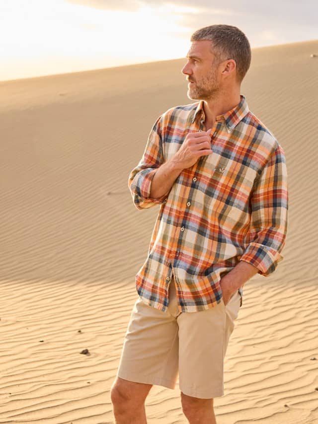 Ein Mann steht in den Dünen und trägt ein kariertes Hemd, dazu helle Shorts. Ein Mann steht in den Dünen und trägt ein kariertes Hemd, dazu helle Shorts.