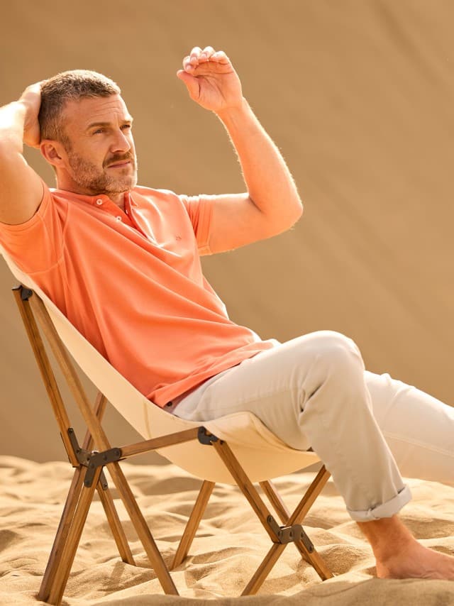 Ein Mann trägt ein Poloshirt in Orange, dazu eine helle Hose und sitzt auf einem Stuhl in den Dünen. Ein Mann trägt ein Poloshirt in Orange, dazu eine helle Hose und sitzt auf einem Stuhl in den Dünen.