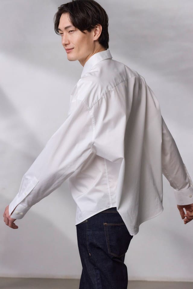 Ein Mann trägt ein weißes Hemd und dunkle Jeans und geht vor einem neutralen Hintergrund.