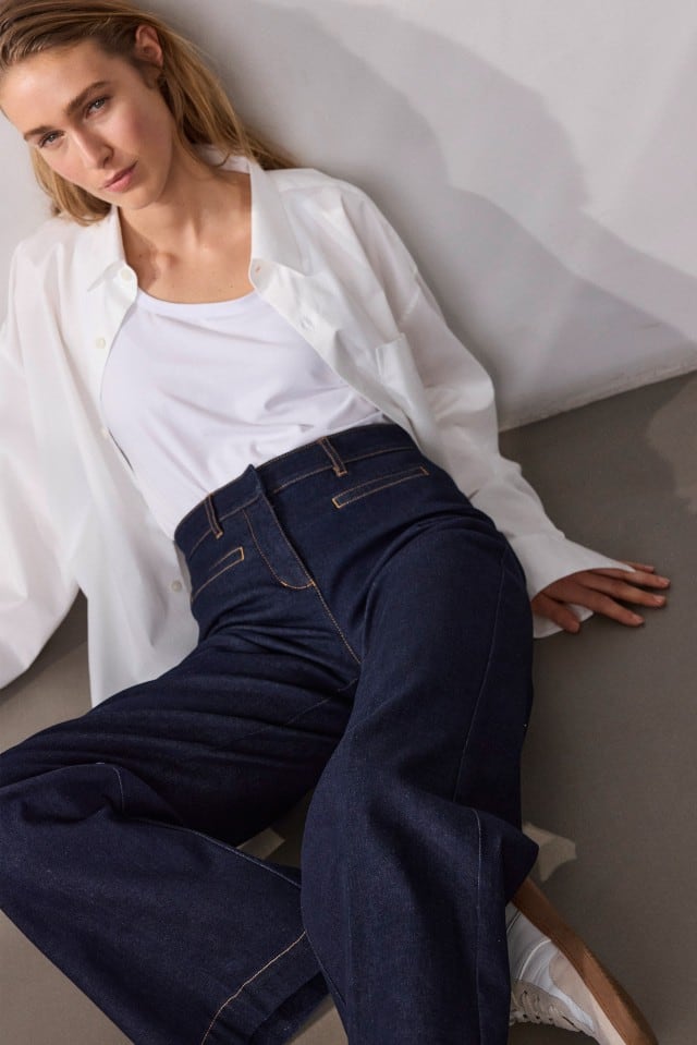 Eine Frau sitzt am Boden und trägt ein weißes Oberteil, ein offenes weißes Hemd sowie dunkelblaue Jeans.