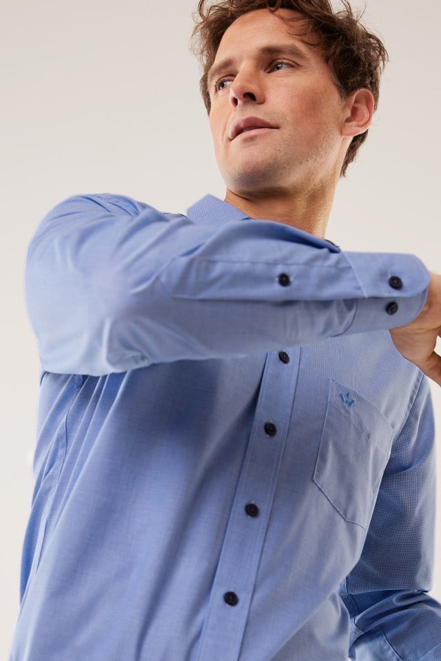 Ein Mann trägt ein hellblaues Hemd und hebt den Arm leicht an, sodass der Fokus auf Ärmel und Knopfleiste liegt.
