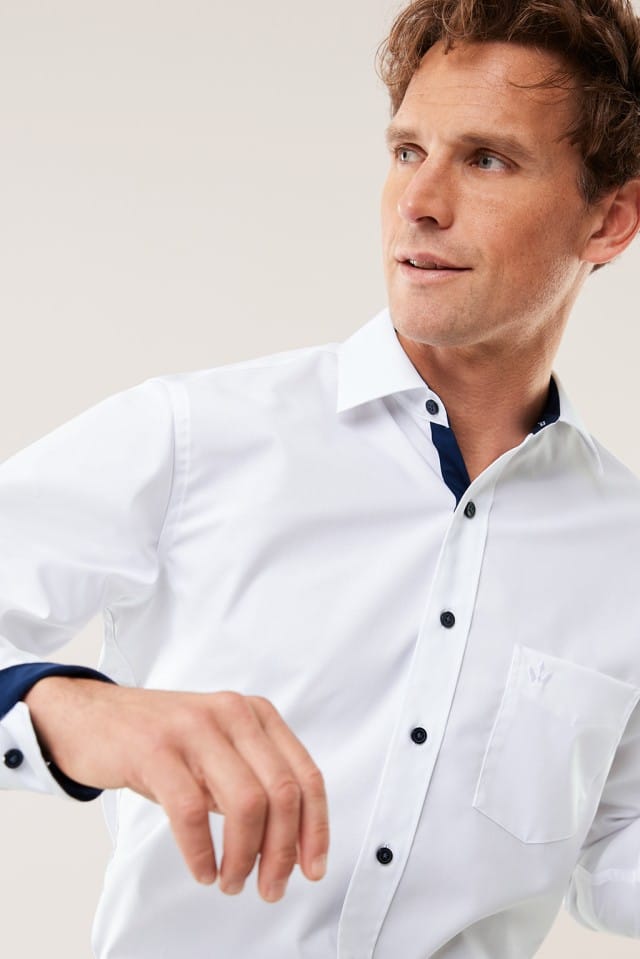 Ein Mann trägt ein weißes Hemd mit dunklen Knöpfen und bewegt leicht den Arm, während er zur Seite blickt.