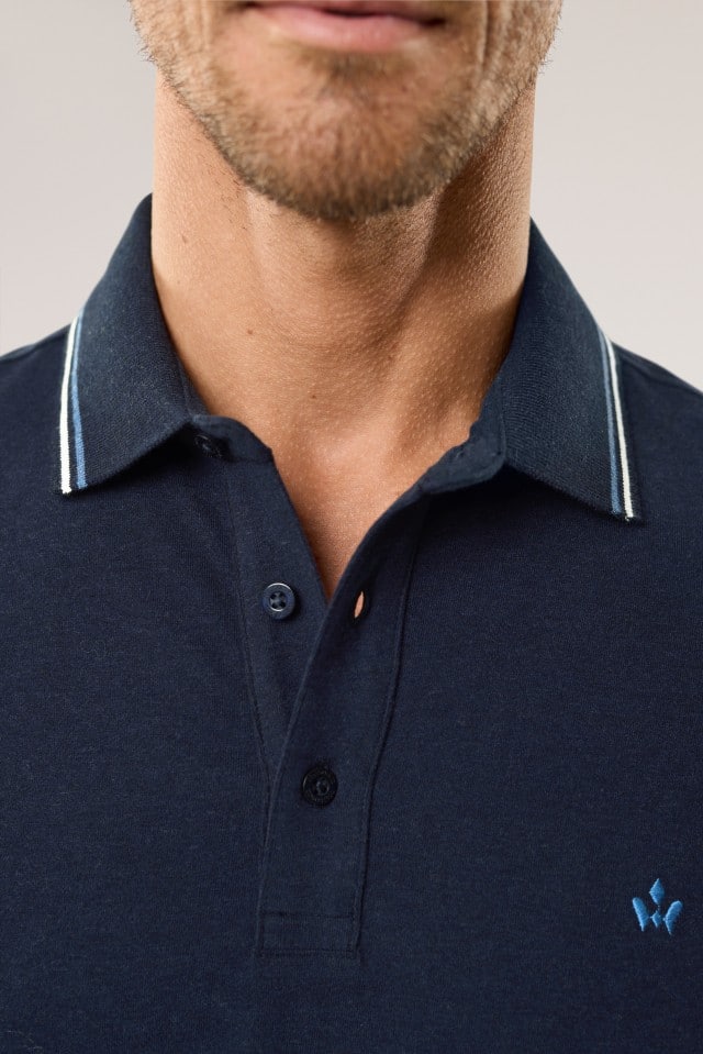 Ein Mann trägt ein dunkelblaues Poloshirt mit feinem, hellen Streifendetail am Kragen und einer dezenten, blauen Stickerei auf der Brust; das Foto zeigt eine Nahaufnahme von Hals und Oberteil.