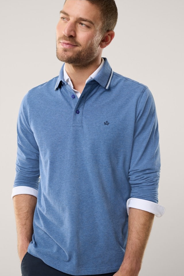 Ein Mann mit kurzem Haar und Bart trägt ein langärmliges, hellblaues Poloshirt mit feiner Kragenkante und aufgesticktem Logo auf der Brust, unter dem ein weißes Hemd leicht hervorblitzt.