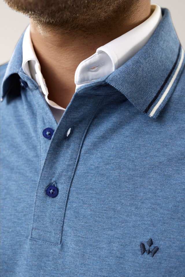 Ein Mann trägt ein hellblaues Poloshirt mit dunkelblauen Knöpfen, feinen Streifendetails am Kragen und einer kleinen Stickerei auf der Brust; unter dem Shirt ist ein weißes Hemd mit klassischem Kragen sichtbar.