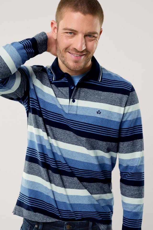 Ein Mann mit kurzem Haar und Bart trägt ein langärmliges Poloshirt mit breiten Querstreifen in verschiedenen Blautönen sowie Grau und Weiß und lächelt, während er eine Hand locker in den Nacken legt.