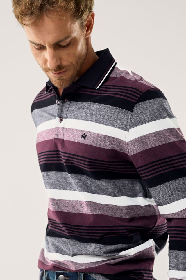 Ein Mann mit kurzem Haar und Bart trägt ein langärmliges Poloshirt mit breiten Querstreifen in Grau-, Weiß-, Bordeaux- und Schwarztönen sowie einem dunklen Kragen mit feiner heller Kante.