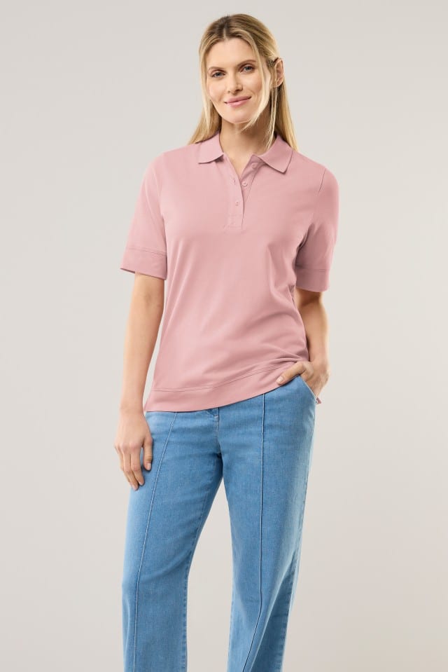 Eine Frau steht vor hellem Hintergrund und trägt ein rosafarbenes Poloshirt mit kurzen Ärmeln sowie eine hellblaue Jeans. Sie lächelt leicht und hat eine Hand in der Hosentasche.