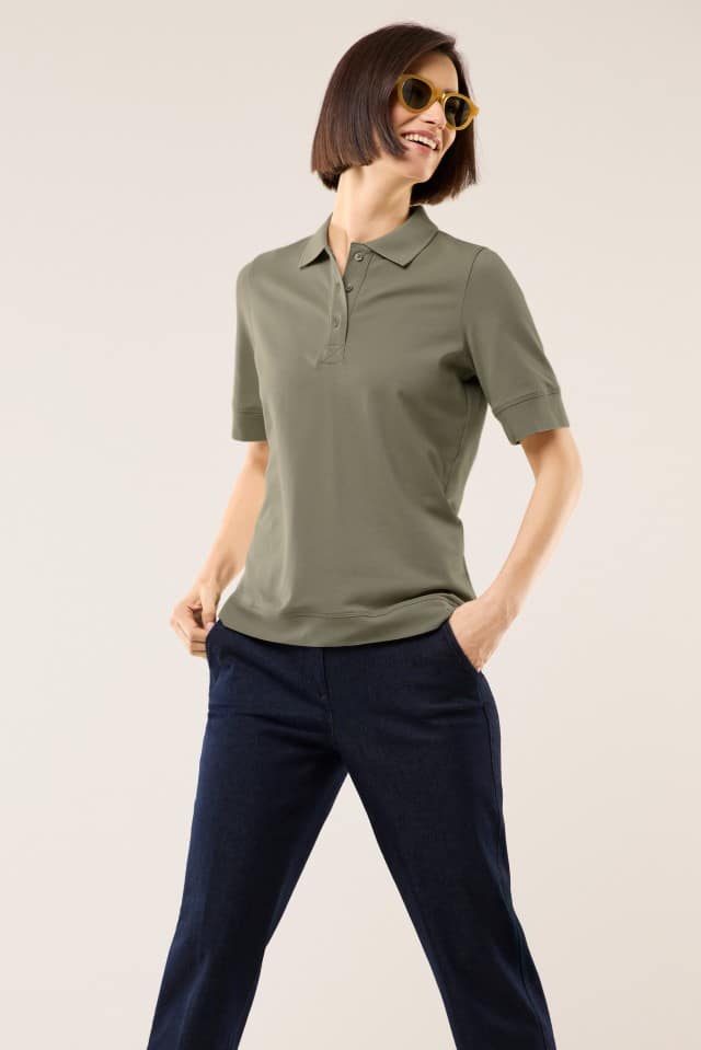 Eine Frau steht vor hellem Hintergrund und trägt ein kurzärmeliges, olivgrünes Poloshirt sowie eine dunkelblaue Hose. Sie lächelt und hat die Hände lässig in den Hosentaschen.