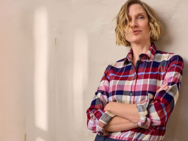 Eine Person steht mit verschränkten Armen und trägt ein kariertes Hemd in Rot-, Blau- und Beigetönen. Eine Person steht mit verschränkten Armen und trägt ein kariertes Hemd in Rot-, Blau- und Beigetönen.