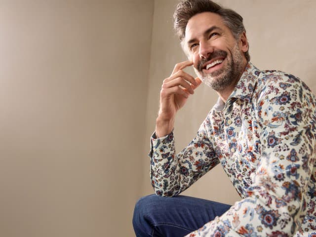 Eine Person sitzt lächelnd und trägt ein helles Hemd mit buntem Blumenmuster, kombiniert mit einer blauen Jeans Eine Person sitzt lächelnd und trägt ein helles Hemd mit buntem Blumenmuster, kombiniert mit einer blauen Jeans