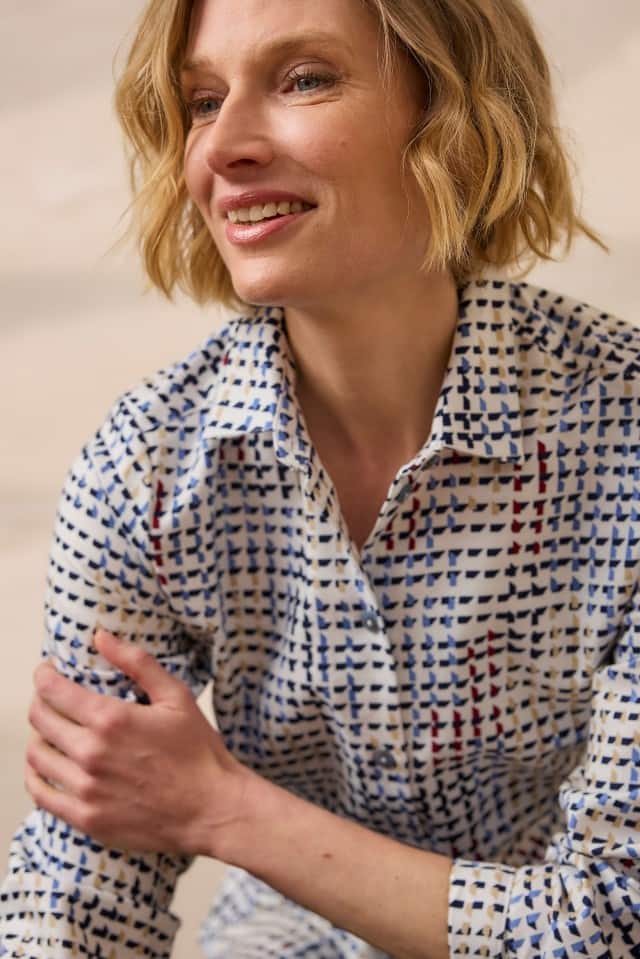 Eine Frau mit blondem, welligem Haar trägt ein gemustertes Hemd in Blau-, Weiß- und Rottönen und lächelt leicht, während sie ihren Arm berührt.