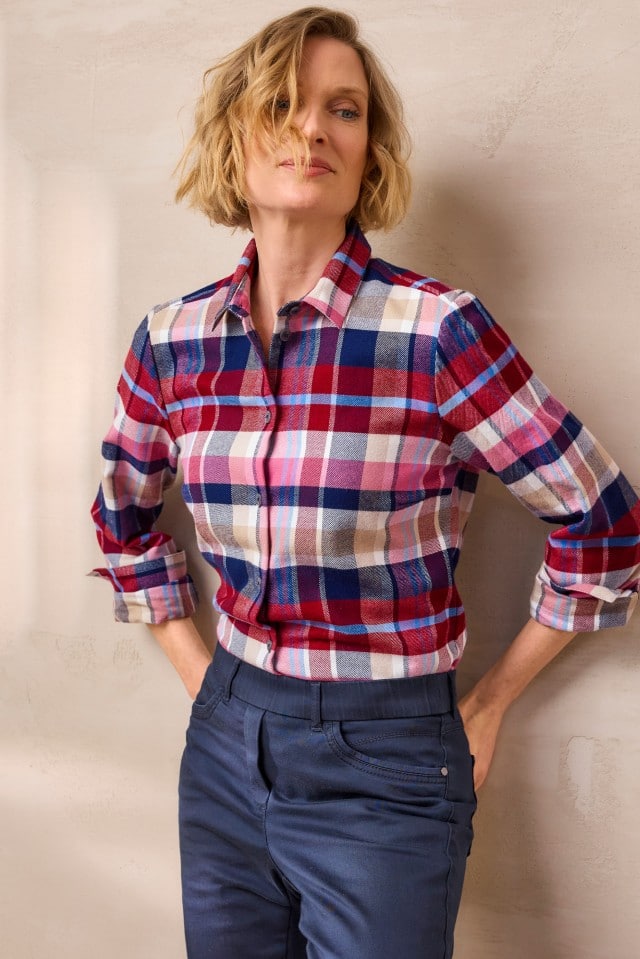 Eine Frau mit blondem, welligem Haar trägt ein kariertes Hemd in Rot-, Blau- und Beigetönen sowie eine dunkelblaue Hose und steht mit den Händen in den Taschen vor einer hellen Wand.