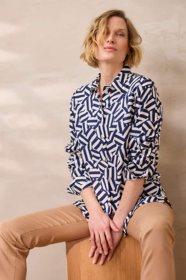 Eine Frau mit blondem, welligem Haar trägt ein weiß-blau gemustertes Hemd mit geometrischem Design und beigefarbene Hose und sitzt mit locker verschränkten Händen auf einem Holzhocker vor einer hellen Wand.