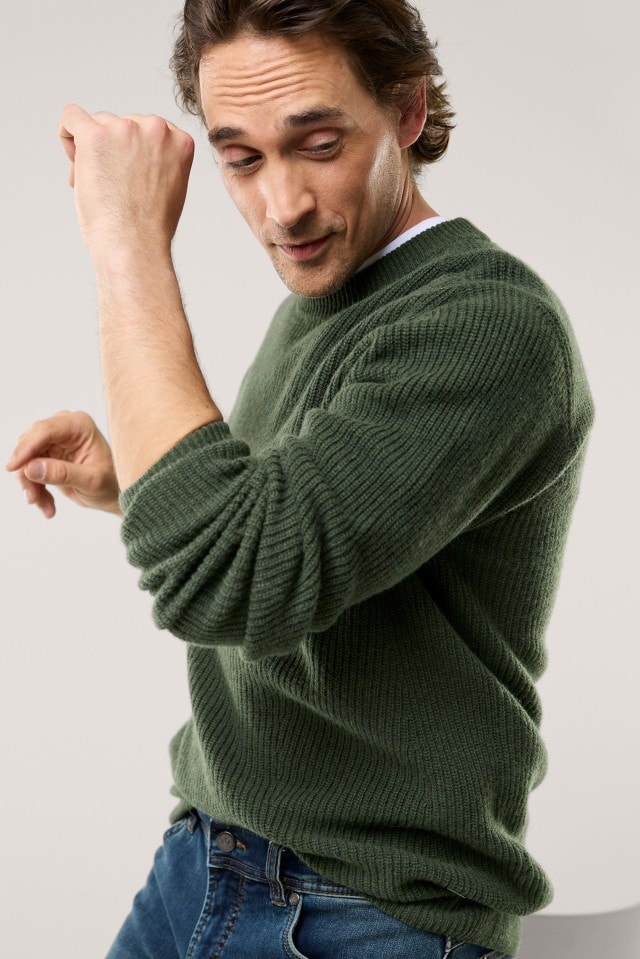 Ein Mann trägt einen dunkelgrün gestrickten Pullover und eine blaue Jeans und bewegt sich in lockerer Pose.