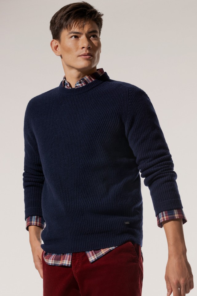 Ein Mann trägt einen dunkelblauen, grob gestrickten Pullover über einem karierten Hemd und kombiniert ihn mit einer roten Cordhose.