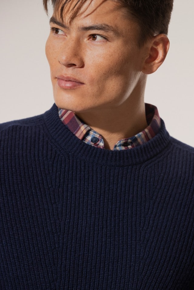 Ein Mann trägt einen dunkelblauen, grob gestrickten Pullover über einem karierten Hemd und blickt zur Seite.