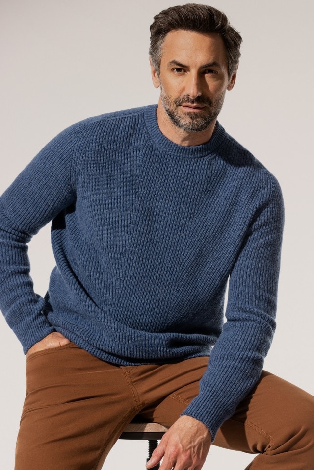 Ein Mann trägt einen blau gestrickten Pullover und kombiniert ihn mit einer braunen Hose.