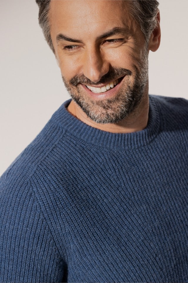 Ein Mann trägt einen grob gestrickten Pullover in Blau und lächelt, während er leicht zur Seite blickt.