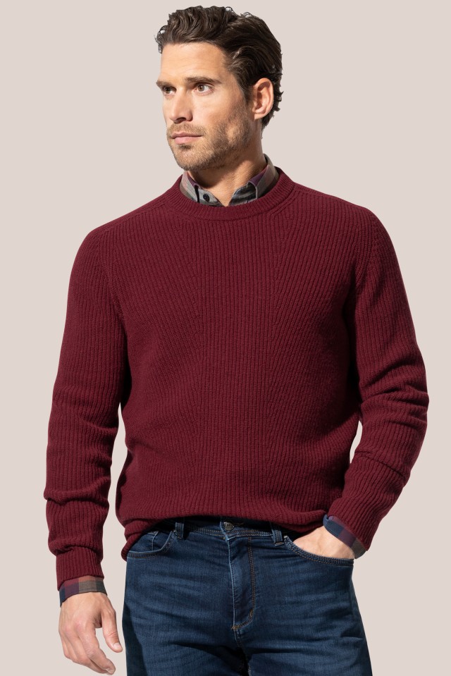 Ein Mann trägt einen grob gestrickten Pullover in Weinrot über einem karierten Hemd und kombiniert ihn mit einer blauen Jeans.