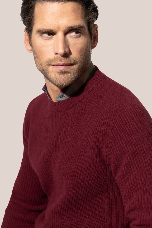 Ein Mann trägt einen weinroten, grob gestrickten Pullover über einem karierten Hemd und blickt zur Seite.