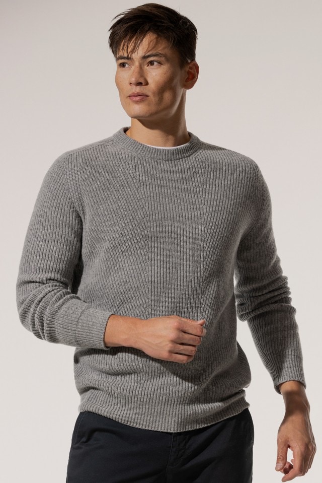 Ein Mann trägt einen hellgrauen, grob gestrickten Pullover über einem weißen Shirt und kombiniert ihn mit einer dunklen Hose.