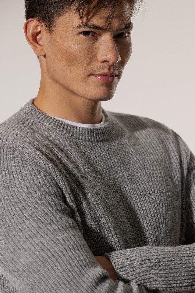Ein Mann trägt einen hellgrauen, grob gestrickten Pullover über einem weißen Shirt und blickt mit verschränkten Armen zur Seite.
