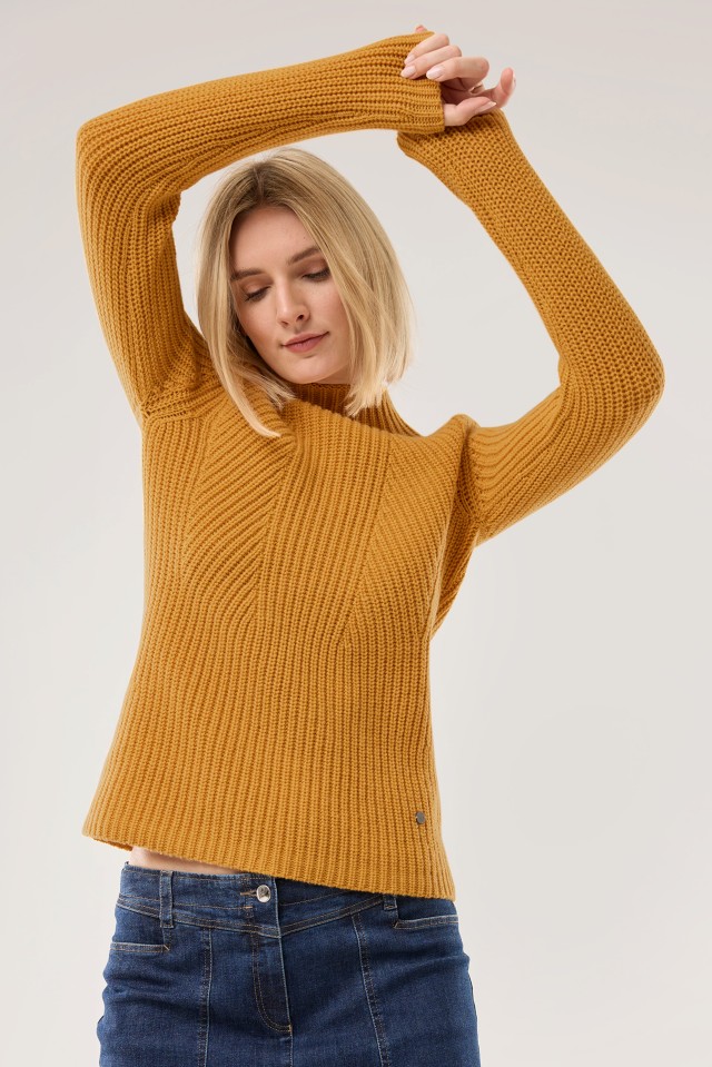 Eine Frau trägt einen safrangelben, grob gestrickten Pullover mit langen Ärmeln und kombiniert ihn mit einer dunkelblauen Jeans.