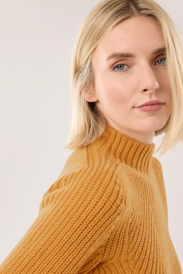 Eine Frau trägt einen safrangelben, grob gestrickten Pullover mit Stehkragen und blickt leicht zur Seite.