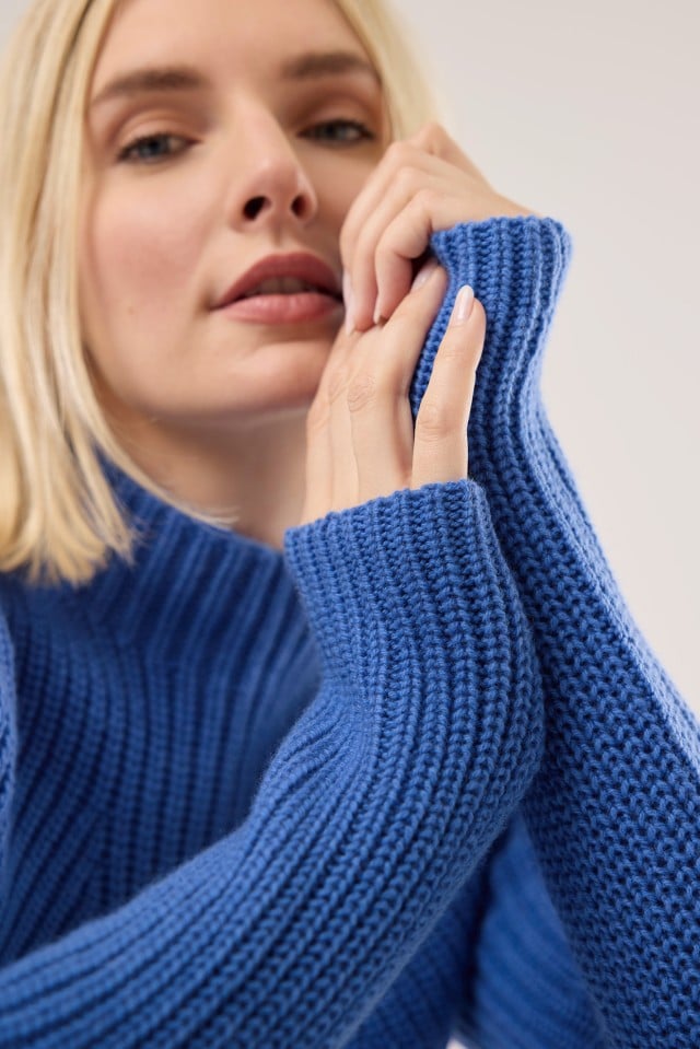 Eine Frau trägt einen blau gestrickten Pullover und hält die langen Ärmel mit beiden Händen nah an ihr Gesicht.