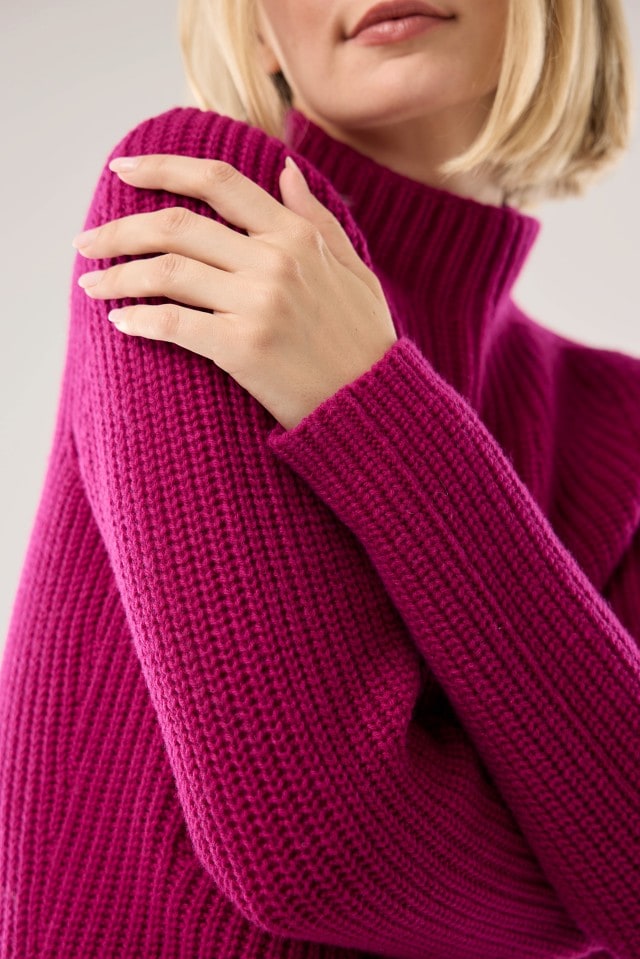 Eine Frau trägt einen grob gestrickten Pullover in kräftigem Beere und legt eine Hand auf ihre Schulter.