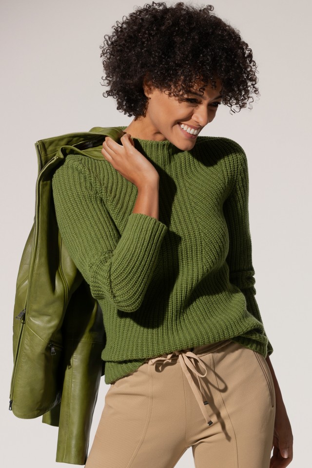 Eine Frau trägt einen grün gestrickten Pullover, hält eine passende grüne Jacke über der Schulter und kombiniert das Outfit mit beigefarbenen Hosen.