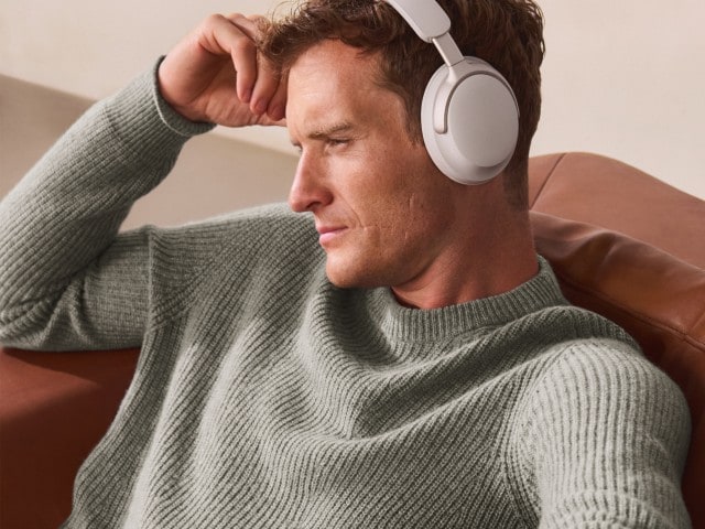 Ein Mann trägt einen hellgrauen, grob gestrickten Pullover und hört mit weißen Kopfhörern Musik, während er entspannt auf einem Sofa sitzt.