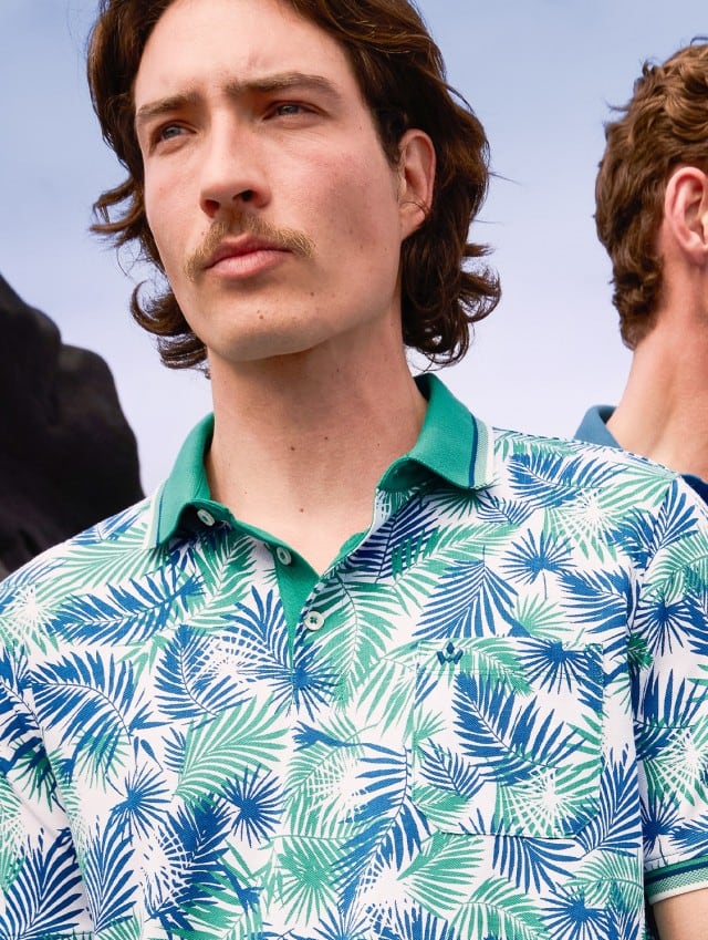 Zwei Männer stehen vor einer felsigen Landschaft: einer im tropisch gemusterten Poloshirt, der andere im hellblauen Poloshirt.