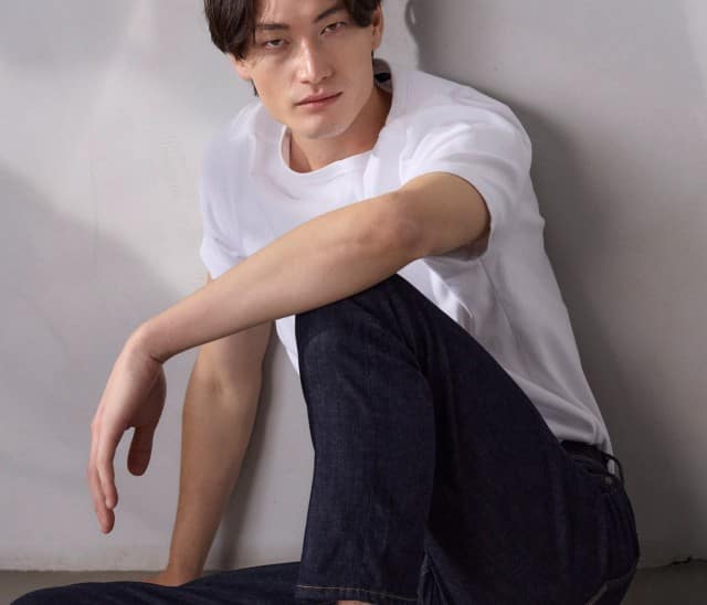 Ein Mann sitzt am Boden vor einer hellen Wand und trägt ein weißes T‑Shirt, dunkle Jeans und weiße Sneaker. Ein Mann sitzt am Boden vor einer hellen Wand und trägt ein weißes T‑Shirt, dunkle Jeans und weiße Sneaker.