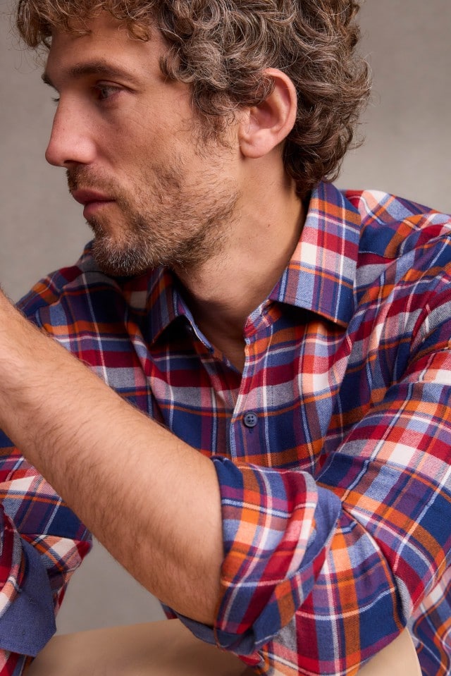 Ein Mann mit lockigem Haar trägt ein bunt kariertes Hemd in Blau-, Rot- und Orangetönen und blickt seitlich nach unten.