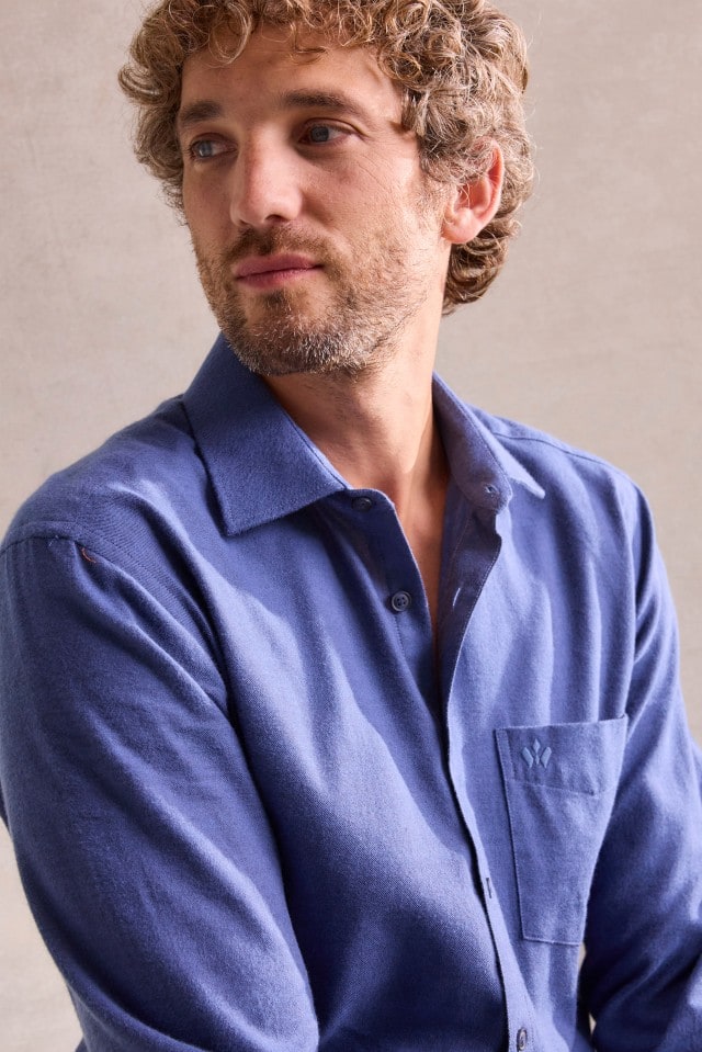 Ein Mann mit lockigem Haar trägt ein blaues Hemd mit Brusttasche und blickt ruhig zur Seite.