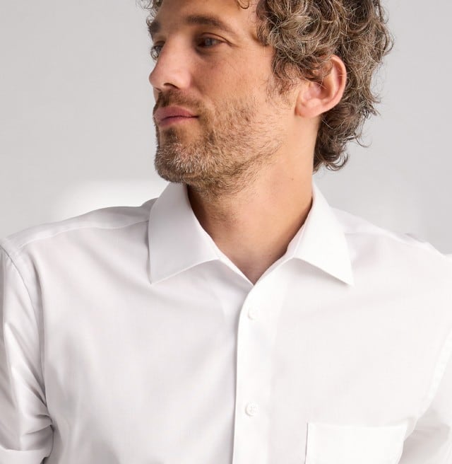 Auf dem Bild ist eine Person mit lockigem, kurzem Haar zu sehen, die ein schlichtes, weißes Hemd trägt und leicht zur Seite blickt.