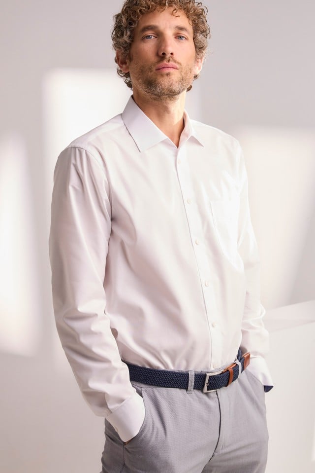 Eine Person steht mit den Händen in den Hosentaschen und trägt ein weißes Hemd, eine graue Hose und einen dunkelblauen Gürtel.