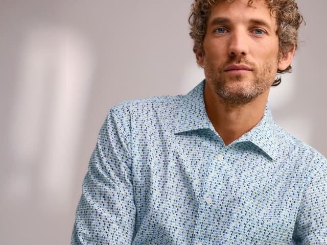 Eine Person mit lockigem Haar trägt ein hell gemustertes Hemd und blickt ruhig in die Kamera.