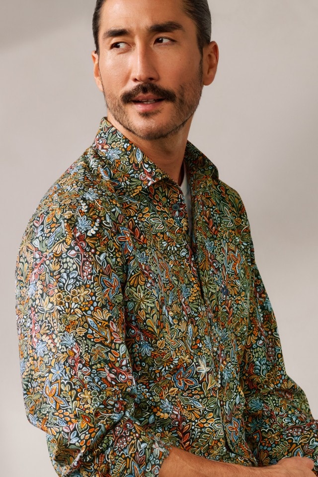 Ein Mann mit Schnurrbart trägt ein bunt gemustertes Hemd mit floralem Design und schaut nach links.