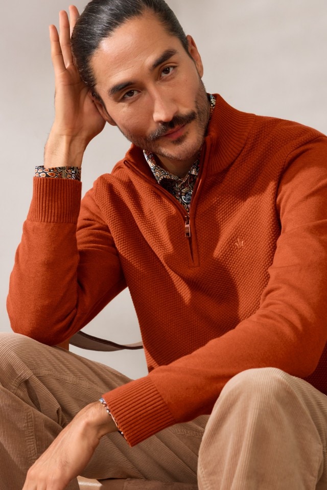 Ein Mann mit zusammengebundenen Haaren und Schnurrbart trägt einen orangefarbenen Strickpullover über einem gemusterten Hemd und sitzt mit der Hand am Kopf.