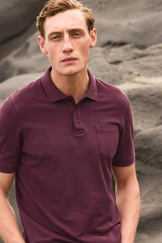 Ein Mann steht vor einer Felslandschaft und trägt ein kurzärmeliges, bordeauxfarbenes Poloshirt mit Brusttasche. Er blickt ruhig in die Kamera.