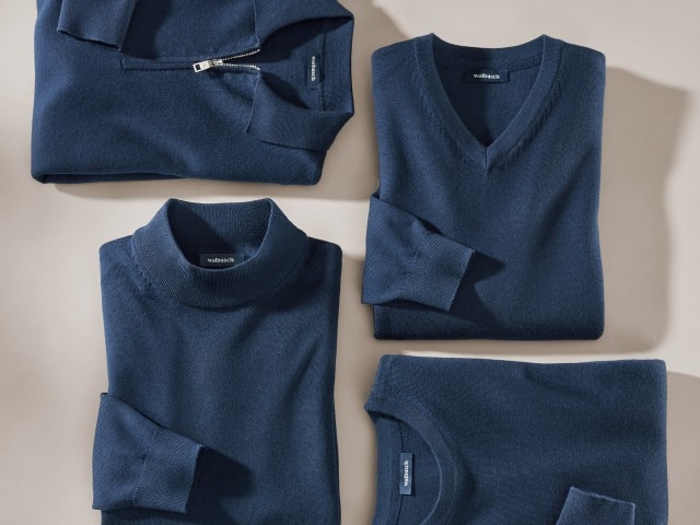 Das Bild zeigt vier gefaltete dunkelblaue Pullover in unterschiedlichen Ausschnitt-Varianten (mit Reißverschlusskragen, V-Ausschnitt, Rollkragen und Rundhals).