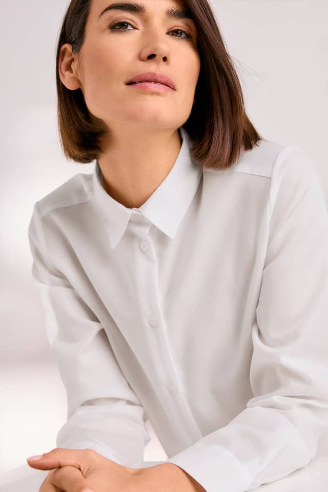 Eine Person mit kinnlangem Haar trägt ein weißes Hemd und blickt ruhig und selbstbewusst in die Kamera.