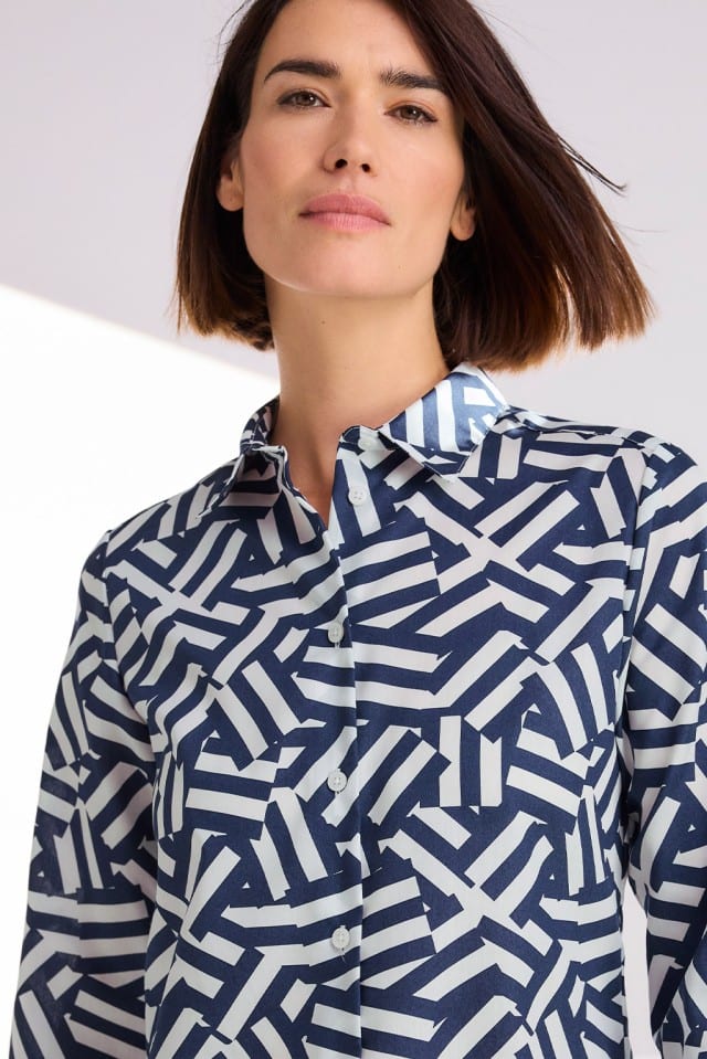 Eine Person mit kinnlangem Haar trägt ein dunkelblau-weiß gemustertes Hemd und blickt ruhig in die Kamera.