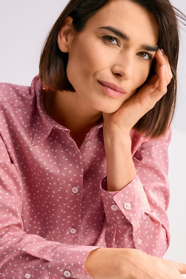 Eine Person mit kinnlangem Haar trägt eine rosafarbene gemusterte Bluse und stützt nachdenklich den Kopf auf die Hand.