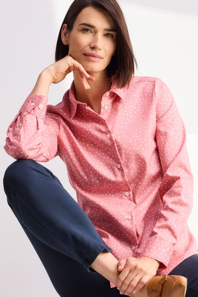 Eine Person mit kinnlangem Haar trägt eine rosafarbene gemusterte Bluse und sitzt entspannt mit angewinkeltem Bein.