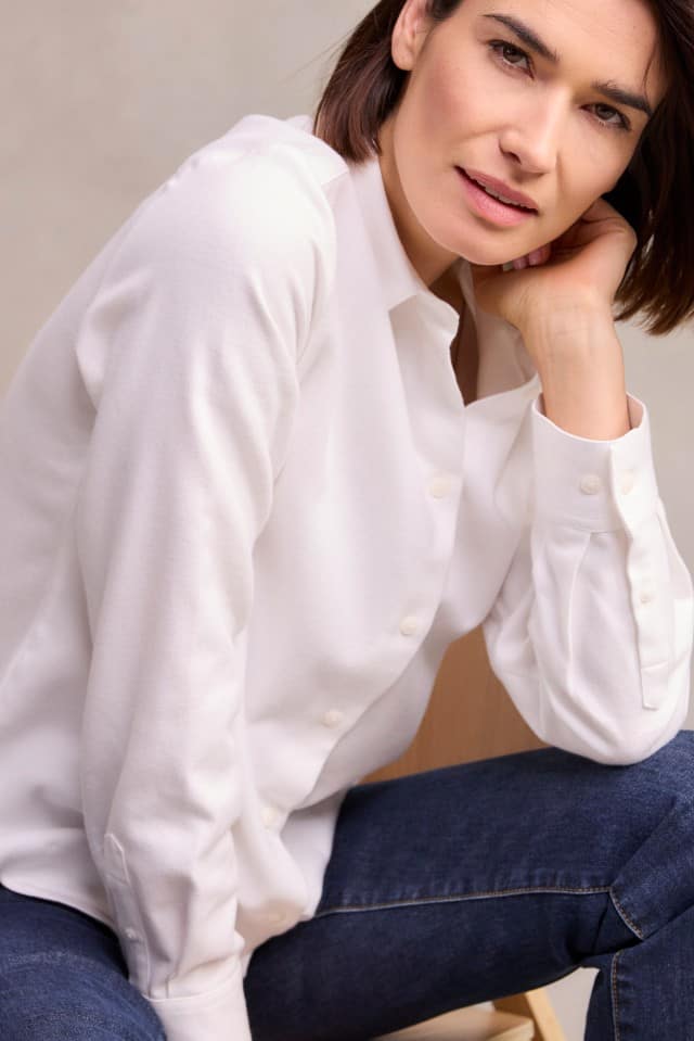 Eine Frau mit schulterlangem, braunem Haar trägt eine weiße Bluse und eine dunkelblaue Jeans, sitzt leicht nach vorn gelehnt auf einem Stuhl und stützt den Kopf auf eine Hand.